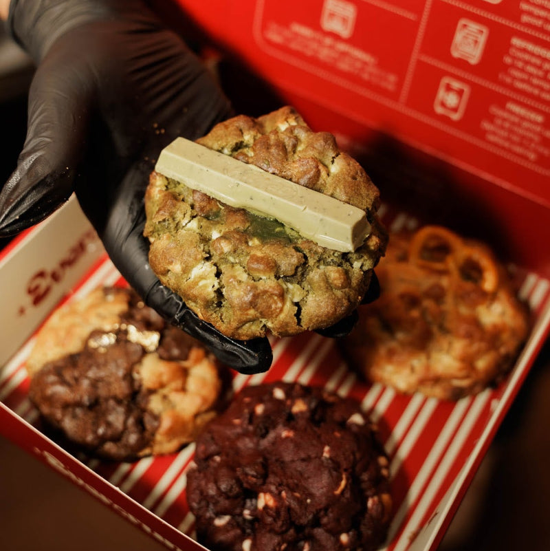 A hand wearing a black glove holds a chocolate chip cookie topped with a bar of chocolate in front of an open red box of cookies. Other chocolate chip cookies are visible inside the box.